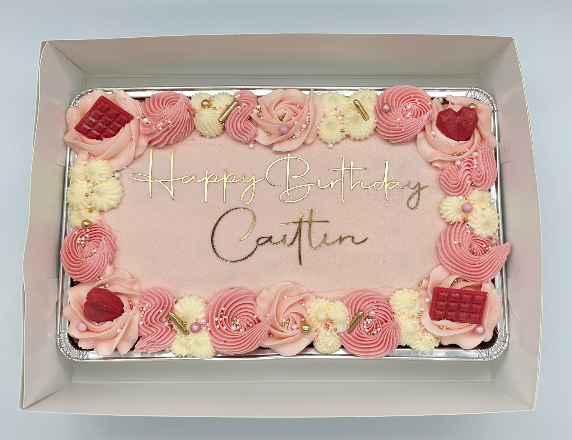 Pink birthday sheet cake with gold lettering, decorated with piped buttercream swirls, edible pearls, gold accents, and chocolate embellishments.